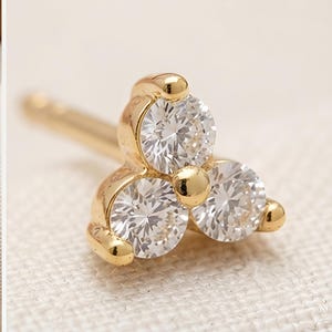 May include: A gold stud earring featuring three round, clear, faceted stones arranged in a triangular cluster. The stones are set in a polished gold metal frame with a small gold bead at the center, and a straight post extends from the back.