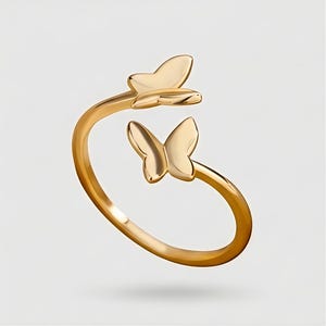 May include: A gold-colored ring featuring two butterfly designs. The ring wraps around the finger with a butterfly at the top and another at the side. The butterflies have detailed wing shapes and are made of a shiny, reflective metal.