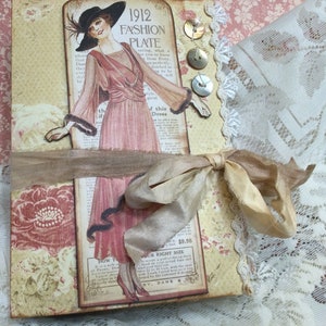 May include: A rectangular journal featuring a vintage 1912 fashion plate illustration of a person in a pink dress and black hat. The cover includes cream floral patterns, three round buttons, white lace trim, and a tan silk ribbon tied in a bow.