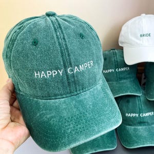Peut inclure: Un groupe de six casquettes de baseball en denim vert avec des broderies blanches qui disent "Happy Camper". Une casquette de baseball blanche avec des broderies vertes qui disent "Bride".