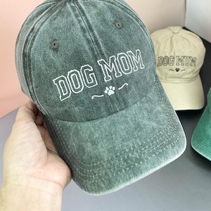 Könnte beinhalten: Drei Baseballkappen in Grün- und Beigetönen. Die Kappen zeigen den gestickten Text "DOG MOM" oder "DOG MUM" in Weiß, zusammen mit einem Pfotenabdruck-Design. Die Kappen sind aus einem abgenutzten Stoff im Vintage-Stil.
