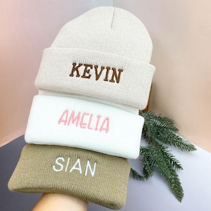 May include: A stack of three personalized knit beanies in cream, white, and tan. Each beanie features an embroidered name: "KEVIN" in brown, "AMELIA" in pink, and "SIAN" in white. The beanies are stacked on top of each other.