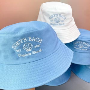May include: A collection of bucket hats in light blue and white. Each hat features white or light blue embroidery with the text ZOEY'S BACH, EST 2026, and Virginia Beach, accompanied by a central seashell graphic on the front crown.