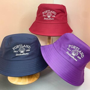 May include: Three bucket hats in dark red, navy blue, and purple. Each hat features white embroidered text reading PORTLAND, JULY 2026, and Bridesthems, centered above a shell graphic. The hats have a wide, downward-sloping brim and a flat crown.