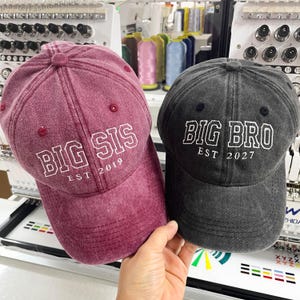 May include: A maroon baseball cap with white embroidered text reading BIG SIS EST 2019 and a charcoal gray baseball cap with white embroidered text reading BIG BRO EST 2027. Both caps feature a distressed fabric finish and a structured crown.