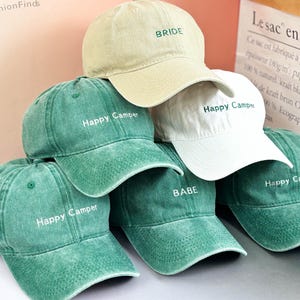 Könnte beinhalten: Fünf grüne und weiße Baseballkappen mit verschiedenen gestickten Texten. Der Text auf den Kappen lautet "Happy Camper", "Babe" und "Bride".
