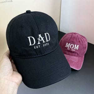 May include: Two baseball caps, one black with "DAD EST. 2025" in white text and one burgundy with "MOM EST. 2025" in white text.