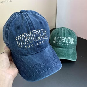 以下が含まれることがあります： 白い刺繍の文字が入ったデニムのベースボールキャップ2つ。青いキャップには「Uncle Est. 2015」、緑のキャップには「Auntie Est. 2025」と書いてあります。