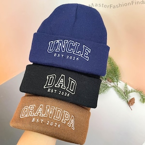 Peut inclure: Trois bonnets en tricot de couleurs bleu marine, noir et marron. Chaque bonnet porte une inscription blanche brodée : "UNCLE EST. 2026", "DAD EST. 2026" et "GRANDPA EST. 2026". Les bonnets sont en tricot doux.