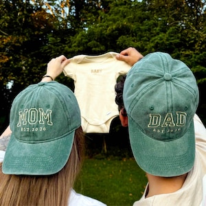 Könnte beinhalten: Zwei grüne Baseballkappen mit den Wörtern "MOM EST. 2026" und "DAD EST. 2026" in Weiß gestickt. Ein cremefarbener Baby-Body mit dem Wort "BABY" wird zwischen den Kappen gehalten. Die Kappen und der Body deuten auf eine Babyankündigung hin.