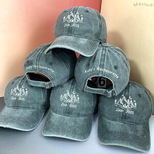 Peut inclure: Plusieurs casquettes de baseball grises empilées. Chaque casquette arbore un motif blanc de montagne et d'arbres avec le texte "Zion 2025". Certaines casquettes portent également l'inscription "Kelly's Bachelorette". Un numéro "26" est visible.