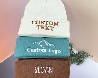 May include: Three stacked knit beanies in white, teal, and brown. The white beanie has "CUSTOM TEXT" embroidered in brown. The teal beanie features a mountain logo and "Custom Logo" in white. The brown beanie displays "SLOAN" in white.