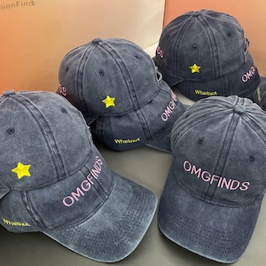 May include: A collection of dark grey baseball caps, each embroidered with the pink text "OMGFINDS". They also feature a yellow star and the word "Whatnot" in yellow. The caps are casually stacked.