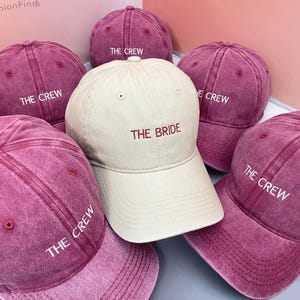 Könnte beinhalten: Eine Auswahl an bordeauxfarbenen Baseballkappen, darunter eine beige Kappe. Die beige Kappe ist mit "THE BRIDE" in Rot bestickt, während die bordeauxfarbenen Kappen "THE CREW" in Weiß tragen. Die Kappen sind locker angeordnet.