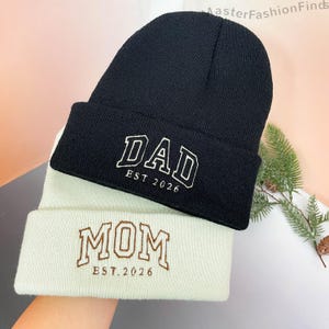 Könnte beinhalten: Zwei Strickmützen, eine schwarze und eine cremefarbene. Die schwarze Mütze hat die weiße Stickerei "DAD EST. 2026". Die cremefarbene Mütze hat die braune Stickerei "MOM EST. 2026". Beide Mützen sind am Rand gefaltet.