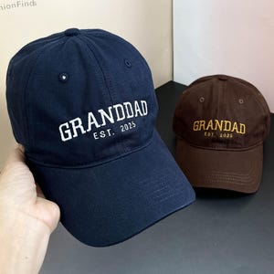 Może przedstawiać: Dwie czapki bejsbolowe, jedna granatowa z białym haftem, na którym widnieje napis "GRANDDAD EST. 2025", a druga brązowa z haftem złotym, na którym widnieje napis "GRANDDAD EST. 2025".