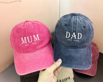 May include: Two denim baseball caps with adjustable straps. One cap is pink with the word "MUM" and "EST. 2025" embroidered in white. The other cap is blue with the word "DAD" and "EST. 2025" embroidered in white.
