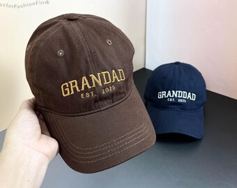 May include: Two baseball caps, one brown and one navy blue, with the word "GRANDDAD" embroidered on the front. The brown cap has gold embroidery and the navy blue cap has white embroidery. Both caps have the text "EST. 2025" embroidered below the word "GRANDDAD".