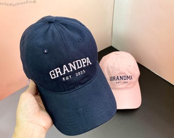 May include: Two baseball caps, one navy blue with "GRANDPA EST. 2025" embroidered in pink, and one pink with "GRANDMA EST. 2025" embroidered in navy blue.