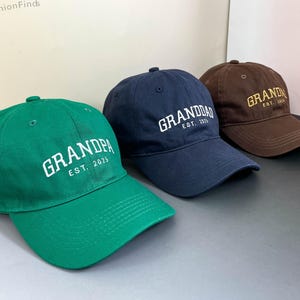 Może przedstawiać: Trzy czapki bejsbolowe w kolorze zielonym, granatowym i brązowym. Każda czapka ma wyhaftowane słowo "GRANDPA" lub "GRANDDAD" w kolorze białym lub złotym, z napisem "EST. 2025" poniżej. Czapki są wykonane z miękkiej tkaniny i mają zakrzywiony daszek.