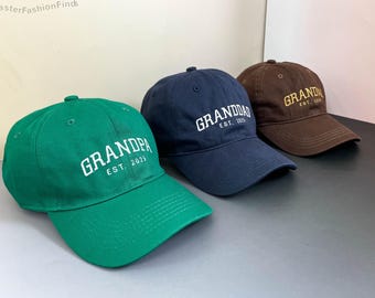 May include: Three baseball caps in green, navy blue, and brown. Each cap has the word "GRANDPA" or "GRANDDAD" embroidered in white or gold, with "EST. 2025" below. The caps are made of a soft fabric and have a curved brim.