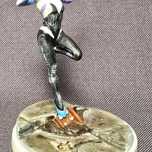 May include: A figurine of a superheroine in a dynamic pose, wearing a black and white suit with a hood. The figure stands on a circular base with a textured surface. The superheroine's shoes are a bright blue color.