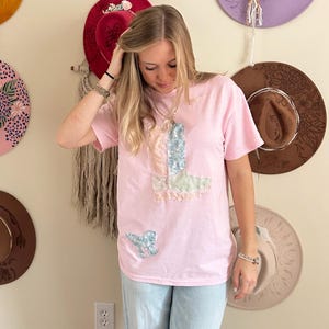 May include: A light pink t-shirt with a patchwork design featuring a boot and butterfly. The shirt is paired with light blue jeans. Various hats in different colors and styles are displayed on the wall behind the person.