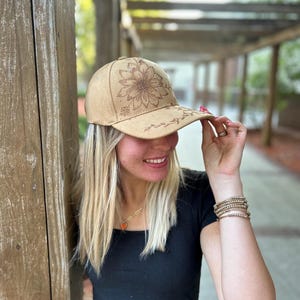 May include: A brown suede baseball cap with a floral design. The cap has a curved brim and an adjustable strap.