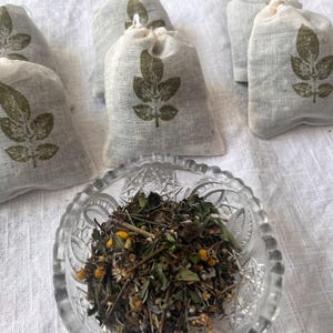 May include: Several small, light-colored fabric bags with a green leaf design are arranged on a white surface. A clear, decorative glass bowl filled with dried herbs and flowers sits in the foreground. The bags appear to be filled with herbs.