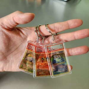 May include: Three Pokémon card keyrings featuring Charizard, Blastoise, and Venusaur. Each card is encased in a clear plastic holder and attached to a silver keyring. The cards have colourful illustrations and text.