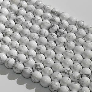 May include: Close-up of a collection of white howlite gemstone beads. The beads are round, with a polished finish and gray marbling patterns. The beads are arranged in rows, creating a visually appealing texture.