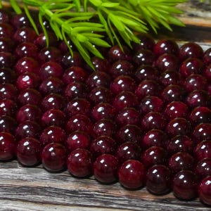May include: A close-up shot of numerous polished, spherical, deep red beads. The beads are tightly packed, reflecting light. Green sprigs are in the background, providing a natural contrast. These beads are likely for jewellery making.