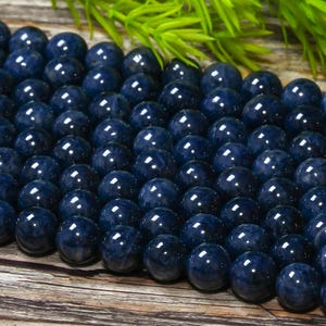 Op de afbeelding: Een close-up van talrijke donkerblauwe edelsteen kralen met een gepolijste, glanzende afwerking. De kralen zijn rond en reflecteren licht. Groen gebladerte is op de achtergrond, rustend op een houten oppervlak. Geschikt voor het maken van sieraden.
