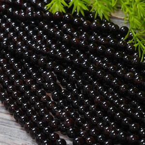 May include: Strands of polished, dark red-brown gemstone beads. The beads are round and uniform in size, with a glossy finish. Green foliage is visible in the upper right corner. The beads are likely for jewelry making.