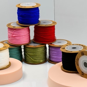 Puede incluir: Una colecci&oacute;n de coloridas bobinas de cord&oacute;n el&aacute;stico en varios colores, incluyendo azul, rosa, rojo, verde, morado, negro y blanco. Las bobinas est&aacute;n apiladas y dispuestas sobre una superficie blanca.