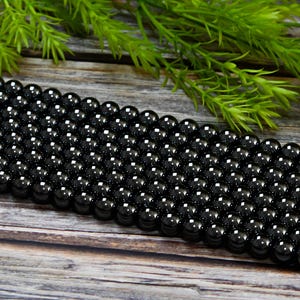 May include: A strand of black obsidian beads, each bead is round and smooth. The beads are arranged in rows on a wooden surface.