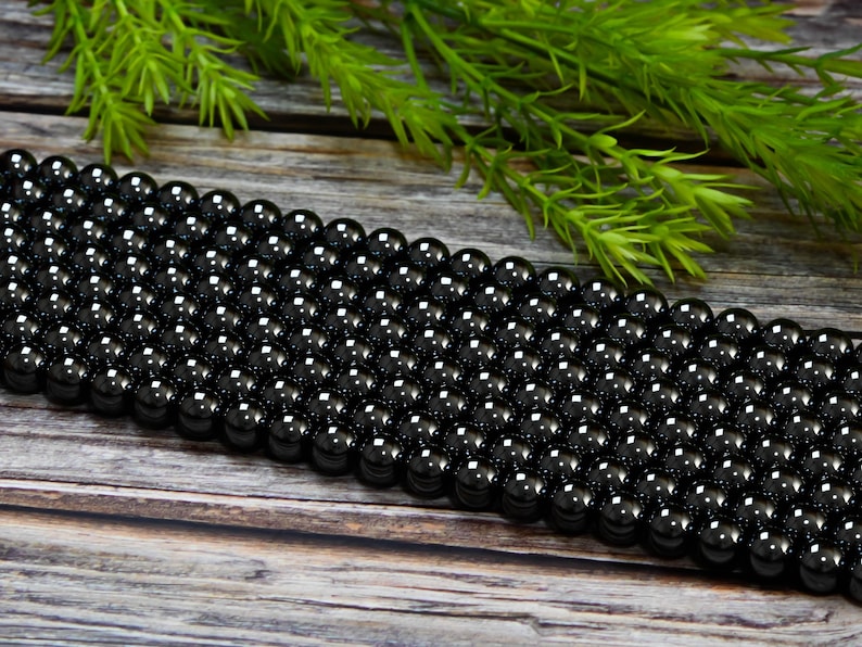 May include: A strand of black obsidian beads, each bead is round and smooth. The beads are arranged in rows on a wooden surface.
