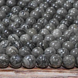 May include: A collection of gray gemstone beads with unique patterns and swirls. The beads are round and polished, varying in shades of gray and black. They are arranged closely together, creating a textured surface.