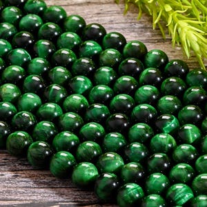 Pode incluir: Um fio de contas de olho de tigre verde. As contas são redondas e lisas, com uma cor verde escura e um brilho sutil.