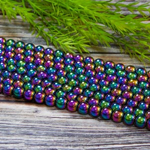 May include: A strand of iridescent rainbow hematite beads. The beads are round and smooth, with a metallic finish. The beads are arranged in a row on a wooden surface.
