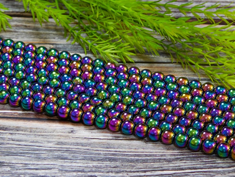 May include: A strand of iridescent rainbow hematite beads. The beads are round and smooth, with a metallic finish. The beads are arranged in a row on a wooden surface.
