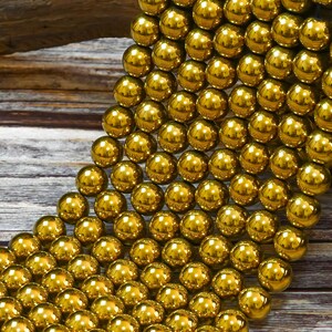 K&ouml;nnte beinhalten: Eine Schnur gl&auml;nzender goldener Perlen, jede Perle ist rund und glatt. Die Perlen sind in einem kaskadierenden Muster auf einer Holzoberfl&auml;che angeordnet.