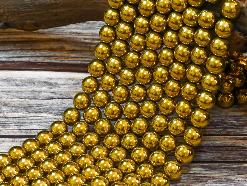 K&ouml;nnte beinhalten: Eine Schnur gl&auml;nzender goldener Perlen, jede Perle ist rund und glatt. Die Perlen sind in einem kaskadierenden Muster auf einer Holzoberfl&auml;che angeordnet.