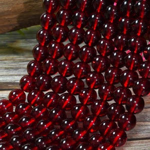 May include: A strand of smooth, spherical beads in a deep, translucent garnet red color. The beads are uniform in size and shape, arranged in a dense, multi-row formation, reflecting light on their polished surfaces with a consistent dark red hue.