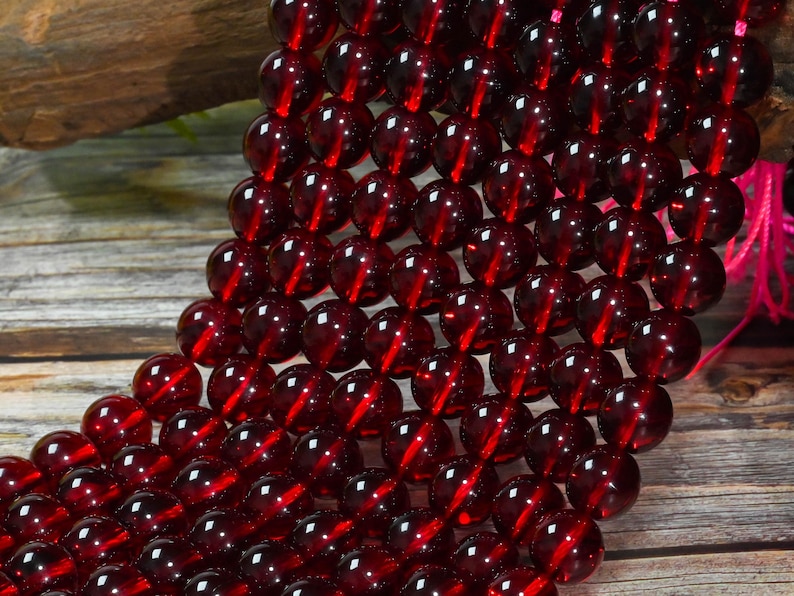 May include: A strand of smooth, spherical beads in a deep, translucent garnet red color. The beads are uniform in size and shape, arranged in a dense, multi-row formation, reflecting light on their polished surfaces with a consistent dark red hue.