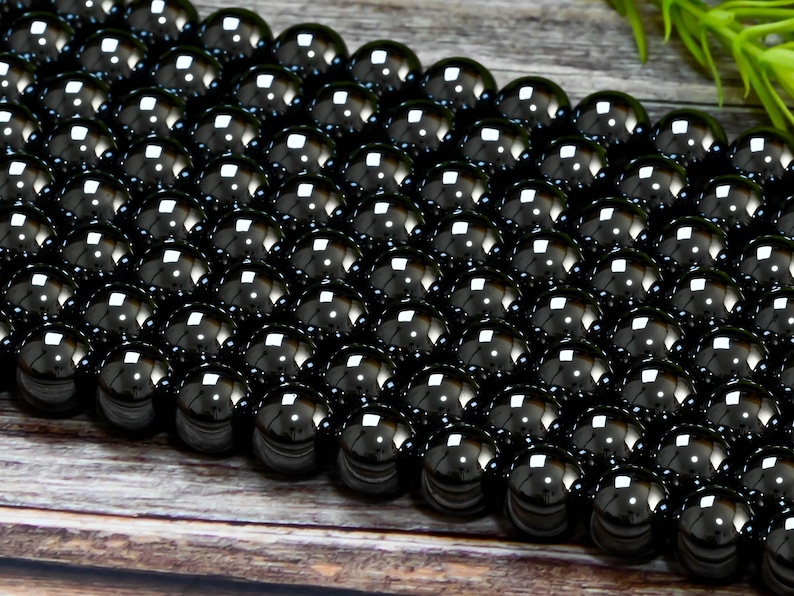 May include: A strand of black obsidian beads, each bead is round and smooth. The beads are arranged in a close-up view, showcasing their glossy, reflective surface.