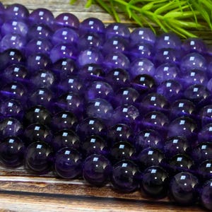 May include: A collection of polished, round amethyst gemstone beads. The beads are a deep purple color, with a glossy finish. The beads are strung together, and are suitable for jewelry making. The background is a wooden surface with green foliage.