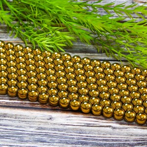 K&ouml;nnte beinhalten: Nahaufnahme einer Reihe gl&auml;nzender goldener Perlen. Die Perlen sind in einem Gittermuster angeordnet und alle gleich gro&szlig;. Die Perlen liegen auf einer Holzoberfl&auml;che.