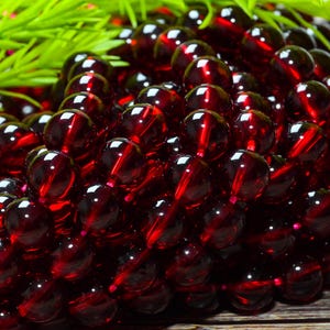 May include: A collection of deep red, spherical garnet beads arranged in multiple strands. Each bead features a smooth, polished, and translucent surface that reflects light, creating a consistent dark crimson appearance across the entire group.