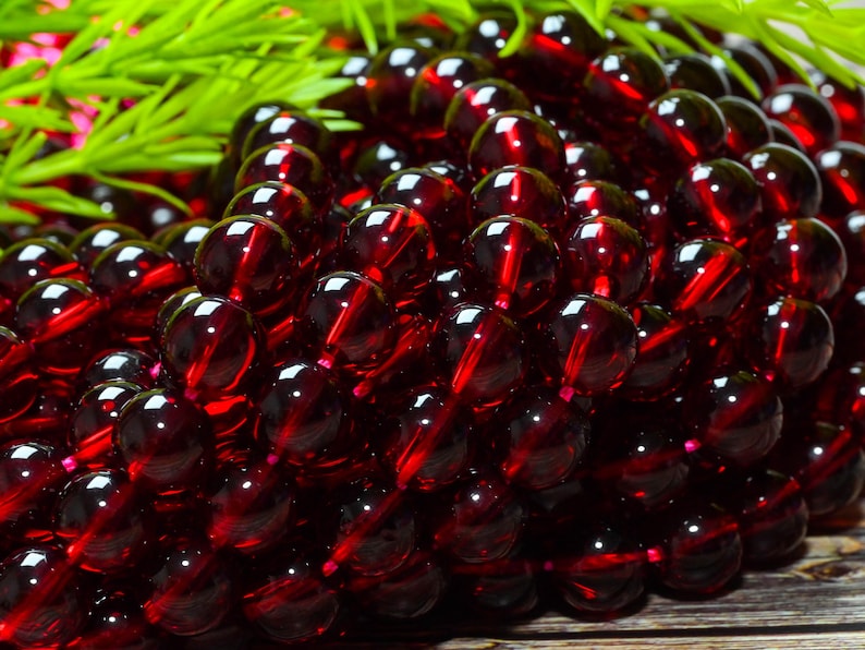 May include: A collection of deep red, spherical garnet beads arranged in multiple strands. Each bead features a smooth, polished, and translucent surface that reflects light, creating a consistent dark crimson appearance across the entire group.